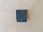 A small dark teal fabric pouch with a flap closure and a bright turquoise snap button, shown on a light beige textured background.