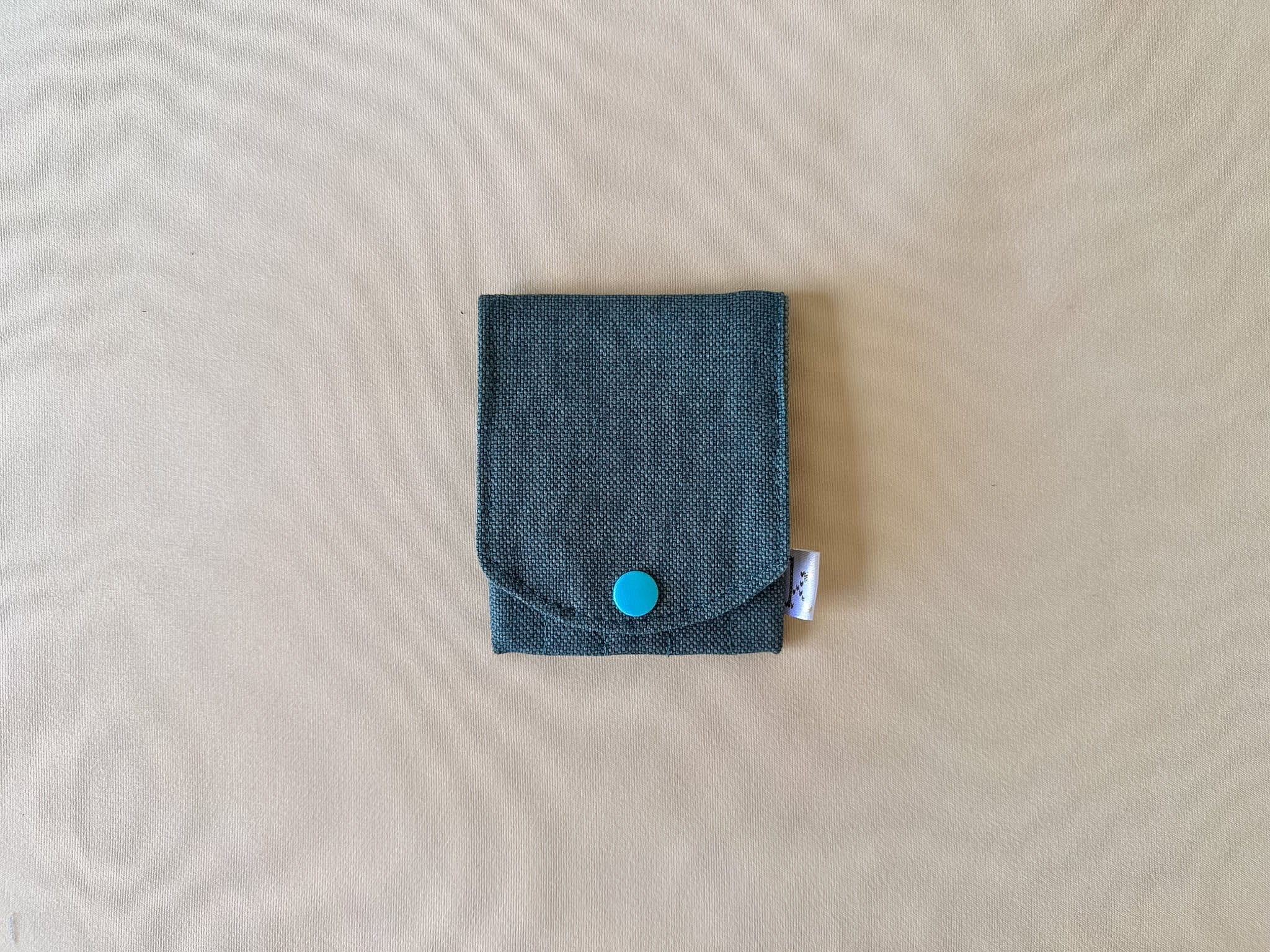 A small dark teal fabric pouch with a flap closure and a bright turquoise snap button, shown on a light beige textured background.